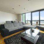 Property for sale by Auction A modern living room in a Liverpool penthouse investment apartment features a large gray sectional sofa and matching ottoman on a black shag rug. A glass-top coffee table sits in front of the sofa, while floor-to-ceiling windows and doors offer an expansive view of the balcony and cityscape beyond. Presented by Palace Auctions