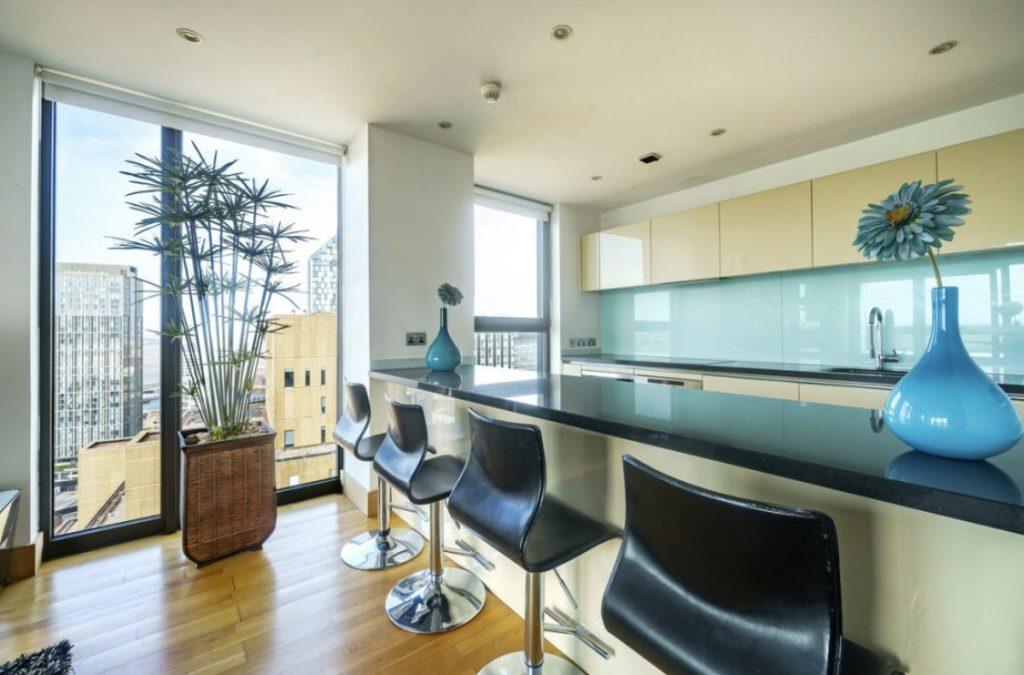 Property for sale by Auction Modern kitchen in a Liverpool penthouse investment apartment features a large island with a black countertop and four matching bar stools. Behind the island, light-colored cabinets, a built-in sink, and large windows provide natural light. Decor includes a tall plant and blue vases with flowers. Presented by Palace Auctions