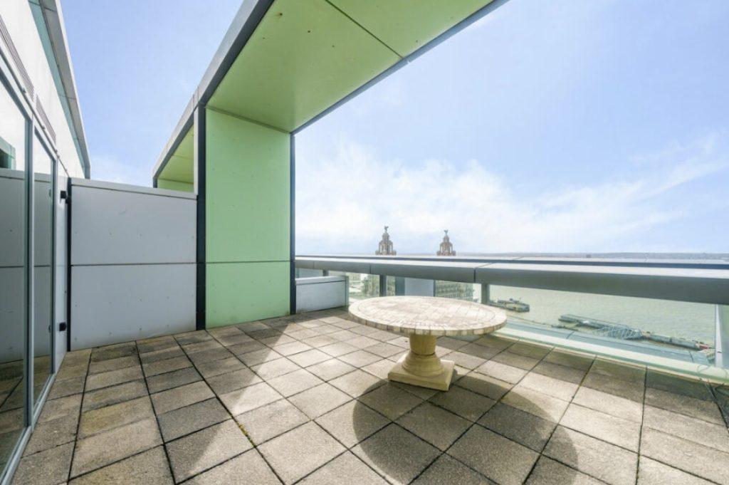 Property for sale by Auction A spacious rooftop terrace in a Liverpool penthouse investment apartment features a green and gray color scheme with a circular stone table at its center. The terrace offers a stunning view of the river and distant buildings under a clear blue sky. Presented by Palace Auctions
