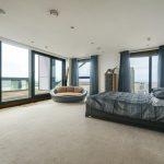 Property for sale by Auction A spacious bedroom in this Liverpool penthouse investment apartment features a large bed with dark bedding, a circular cushioned chair, a tall dresser, and floor-to-ceiling windows offering a panoramic city view. The room is decorated in neutral tones with blue accents and has a door leading to a private balcony. Presented by Palace Auctions