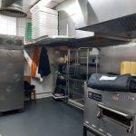 Property for sale by Auction A commercial kitchen with stainless steel appliances, shelves stocked with containers, and pizza boxes stacked high reflects the bustling activity of Pizza Shop 4 U. A person sits on a bench to the left, partially visible. An oven, refrigerator, and cooking hood are prominent in this mixed commercial and residential leasehold investment property. Presented by Palace Auctions