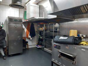 Property for sale by Auction A commercial kitchen with stainless steel appliances, shelves stocked with containers, and pizza boxes stacked high reflects the bustling activity of Pizza Shop 4 U. A person sits on a bench to the left, partially visible. An oven, refrigerator, and cooking hood are prominent in this mixed commercial and residential leasehold investment property. Presented by Palace Auctions