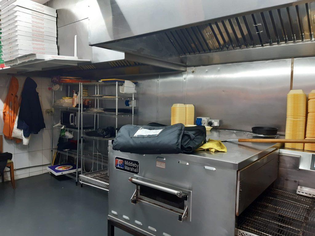 Property for sale by Auction A commercial kitchen in a bustling pizza shop 4 u, featuring a large industrial oven with a bag on top, shelving with pizza boxes, a microwave, and various supplies. The stainless steel walls and large ventilation hoods add to its efficiency. Yellow containers are stacked on a counter in the background. Presented by Palace Auctions