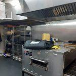 Property for sale by Auction A commercial kitchen in a bustling pizza shop 4 u, featuring a large industrial oven with a bag on top, shelving with pizza boxes, a microwave, and various supplies. The stainless steel walls and large ventilation hoods add to its efficiency. Yellow containers are stacked on a counter in the background. Presented by Palace Auctions