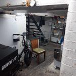 Property for sale by Auction A basement area with white brick walls and various items scattered around, reminiscent of a storage space in a mixed commercial and residential leasehold investment property. There's a bike labeled "BIKE IT" near a white cabinet, a wooden chair with a green seat in the foreground, metal shelves, and a staircase leading to an upper level. Presented by Palace Auctions