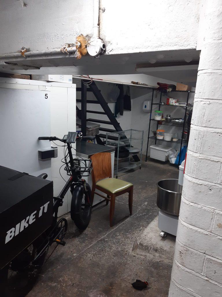 Property for sale by Auction A basement area with white brick walls and various items scattered around, reminiscent of a storage space in a mixed commercial and residential leasehold investment property. There's a bike labeled "BIKE IT" near a white cabinet, a wooden chair with a green seat in the foreground, metal shelves, and a staircase leading to an upper level. Presented by Palace Auctions