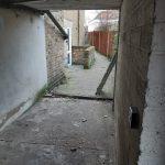 Property for sale by Auction A narrow, semi-enclosed passageway with bare concrete and brick walls leads to an open, paved area. The ground appears old and worn, with scattered debris. In the distance, there's a brick building housing a pizza shop 4 u alongside other mixed commercial and residential leasehold investment properties. Presented by Palace Auctions