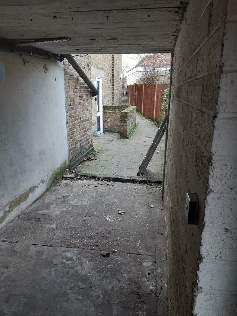 Property for sale by Auction A narrow, semi-enclosed passageway with bare concrete and brick walls leads to an open, paved area. The ground appears old and worn, with scattered debris. In the distance, there's a brick building housing a pizza shop 4 u alongside other mixed commercial and residential leasehold investment properties. Presented by Palace Auctions
