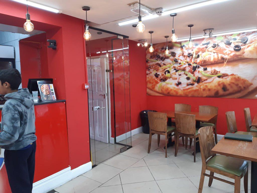 Property for sale by Auction A small pizza shop 4 u with bright red walls, wooden chairs, and tables. The wall features a large, vibrant image of pizzas. Edison bulbs hang from the ceiling. A boy in a hoodie stands near the counter on the left. The décor is clean and modern, making it an attractive mixed commercial and residential leasehold investment property. Presented by Palace Auctions