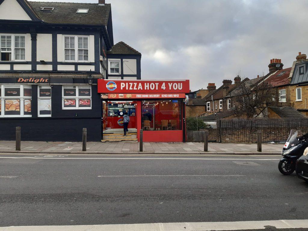Property for sale by Auction A street view of "Pizza Hot 4 You," a pizza shop with a red and white storefront in a mixed commercial and residential area. The building, featuring black trim, is next to homes, making it an attractive leasehold investment property. A man on a motorcycle rides past the shop on the road. Presented by Palace Auctions