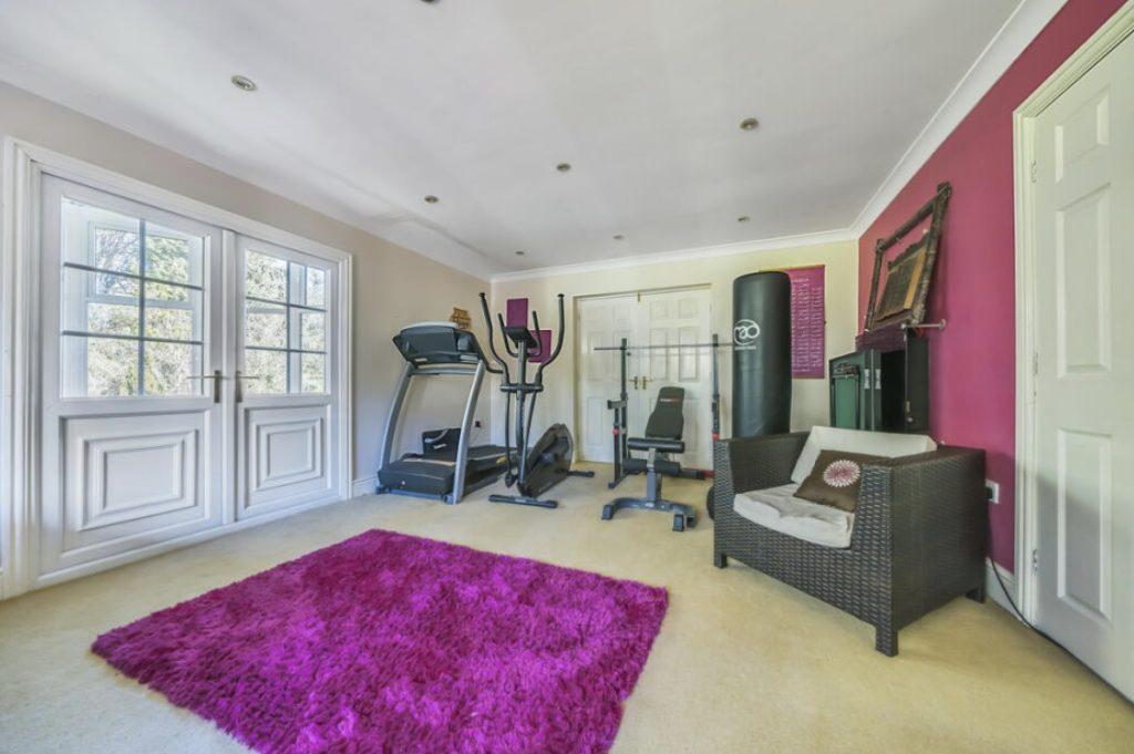 Property for sale by Auction A home gym on Rowan Avenue with exercise equipment including a treadmill, stationary bike, weight bench, and punching bag. The room features a bright pink wall, a white double door with windows, recessed lighting, a purple rug, and a wicker chair with a cushion. Presented by Palace Auctions