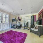 Property for sale by Auction A home gym on Rowan Avenue with exercise equipment including a treadmill, stationary bike, weight bench, and punching bag. The room features a bright pink wall, a white double door with windows, recessed lighting, a purple rug, and a wicker chair with a cushion. Presented by Palace Auctions