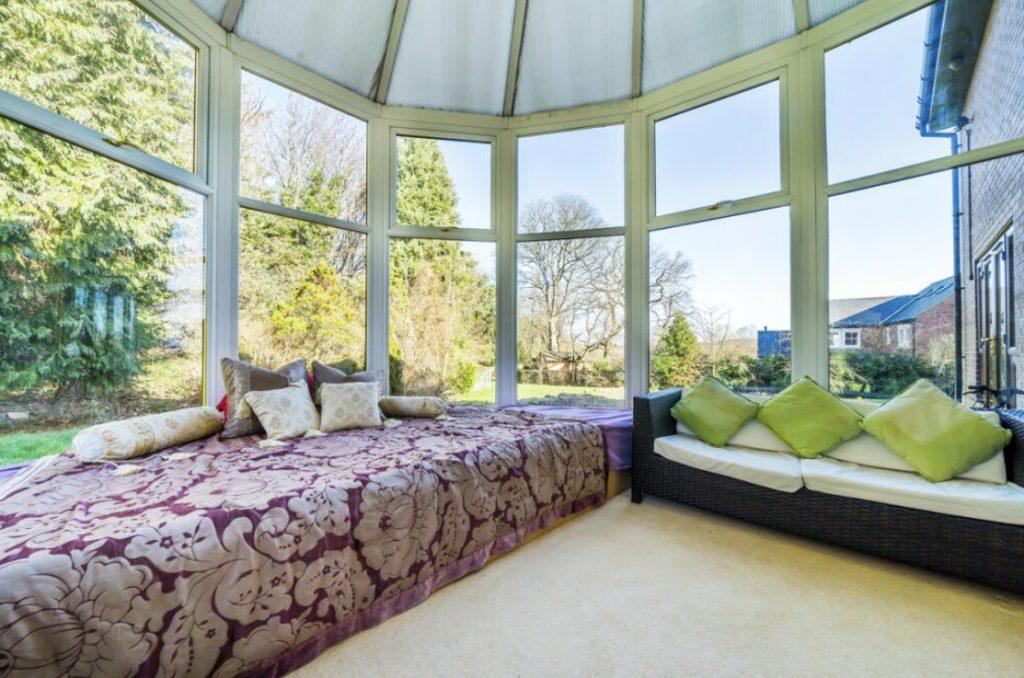 Property for sale by Auction A sunlit conservatory on Rowan Avenue boasts large windows showcasing a garden view. It features a cozy setup with a daybed adorned with floral-patterned bedding and numerous cushions, while a wicker sofa with green and white pillows sits adjacent to the daybed. Presented by Palace Auctions