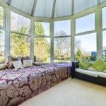 Property for sale by Auction A sunlit conservatory on Rowan Avenue boasts large windows showcasing a garden view. It features a cozy setup with a daybed adorned with floral-patterned bedding and numerous cushions, while a wicker sofa with green and white pillows sits adjacent to the daybed. Presented by Palace Auctions