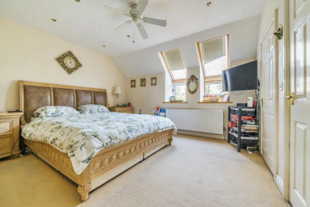 Property for sale by Auction A spacious bedroom on Rowan Avenue features a large, wooden, king-size bed with a floral-patterned bedspread. The room has a beige carpet, an angled ceiling with a ceiling fan, two skylight windows, a wall-mounted TV, a wooden dresser, and a door on the right. Presented by Palace Auctions
