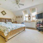 Property for sale by Auction A spacious bedroom on Rowan Avenue features a large, wooden, king-size bed with a floral-patterned bedspread. The room has a beige carpet, an angled ceiling with a ceiling fan, two skylight windows, a wall-mounted TV, a wooden dresser, and a door on the right. Presented by Palace Auctions