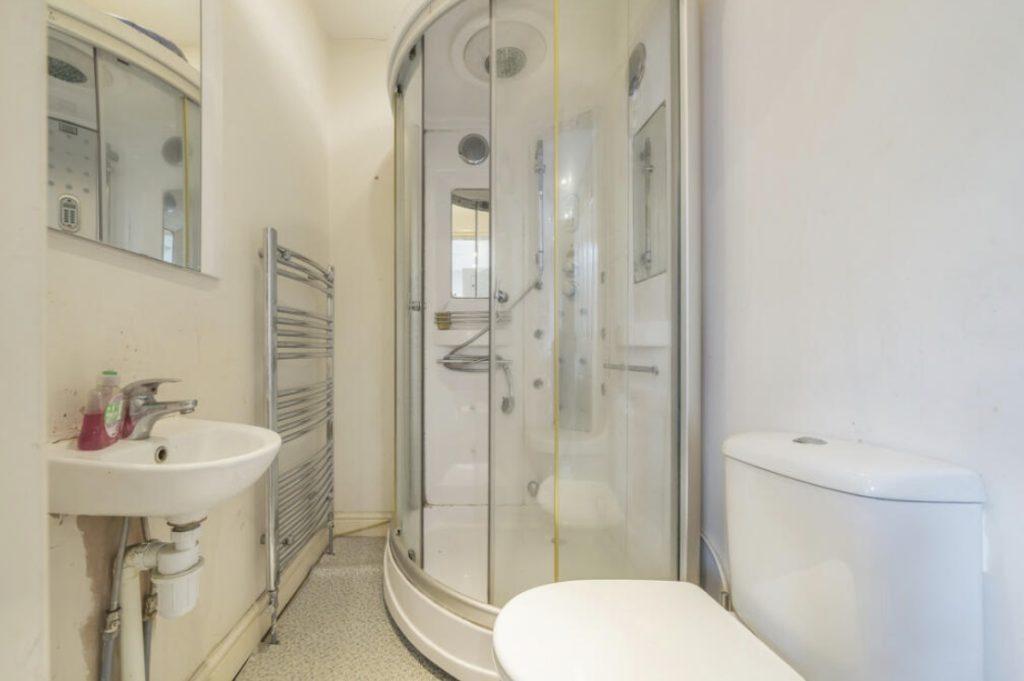 Property for sale by Auction A small bathroom on Rowan Avenue features a white sink on the left with a wall-mounted mirror above it, a silver towel radiator on the left wall, a rounded glass shower enclosure in the center, and a white toilet on the right side against a pale-colored wall. Presented by Palace Auctions