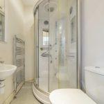 Property for sale by Auction A small bathroom on Rowan Avenue features a white sink on the left with a wall-mounted mirror above it, a silver towel radiator on the left wall, a rounded glass shower enclosure in the center, and a white toilet on the right side against a pale-colored wall. Presented by Palace Auctions