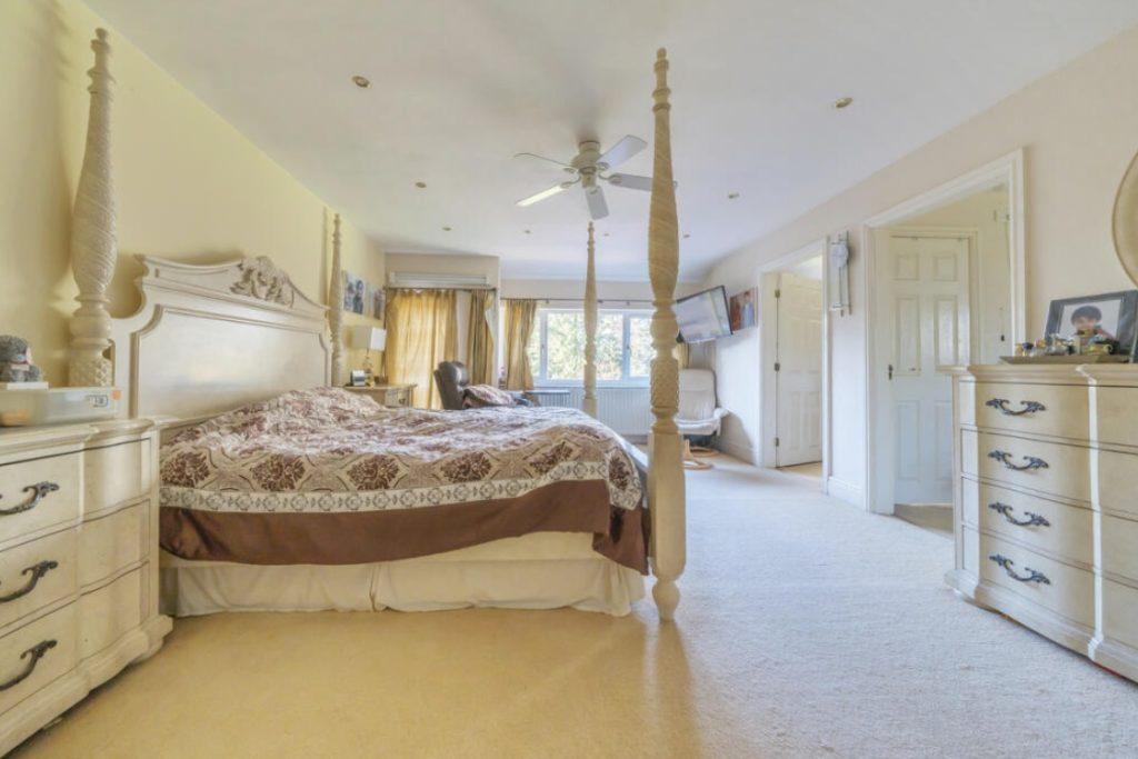 Property for sale by Auction A spacious, well-lit bedroom on Rowan Avenue features cream-colored walls and carpets. It includes a large four-poster bed with a decorative headboard, matching nightstands, and a dresser. There's also a ceiling fan, a window with yellow curtains, and a TV mounted on the wall. Presented by Palace Auctions
