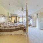 Property for sale by Auction A spacious, well-lit bedroom on Rowan Avenue features cream-colored walls and carpets. It includes a large four-poster bed with a decorative headboard, matching nightstands, and a dresser. There's also a ceiling fan, a window with yellow curtains, and a TV mounted on the wall. Presented by Palace Auctions
