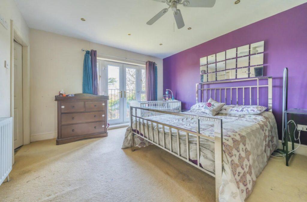 Property for sale by Auction A bedroom on Rowan Avenue with light-colored walls and a purple accent wall. It features a metallic bed with patterned bedding, a ceiling fan, a wooden dresser, and French doors leading to a balcony. The room is bright with light streaming through the doors. Presented by Palace Auctions