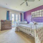 Property for sale by Auction A bedroom on Rowan Avenue with light-colored walls and a purple accent wall. It features a metallic bed with patterned bedding, a ceiling fan, a wooden dresser, and French doors leading to a balcony. The room is bright with light streaming through the doors. Presented by Palace Auctions