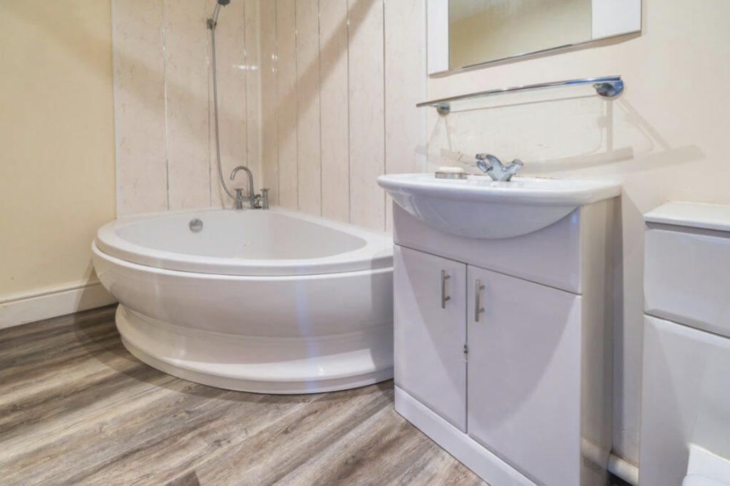 Property for sale by Auction A modern bathroom on Rowan Avenue featuring a round, white bathtub with a wall-mounted showerhead. A white vanity with a sink and mirror is positioned next to the tub. The room has light-colored walls and wood-look vinyl flooring. Presented by Palace Auctions