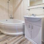 Property for sale by Auction A modern bathroom on Rowan Avenue featuring a round, white bathtub with a wall-mounted showerhead. A white vanity with a sink and mirror is positioned next to the tub. The room has light-colored walls and wood-look vinyl flooring. Presented by Palace Auctions