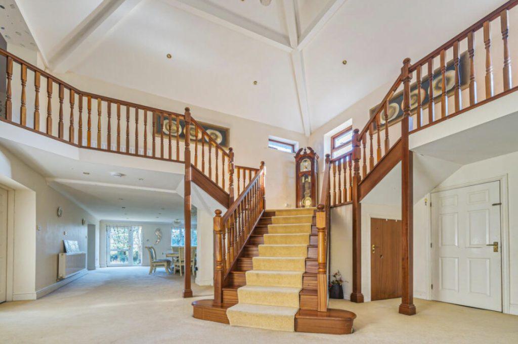 Property for sale by Auction A grand foyer on Rowan Avenue features a wide, carpeted staircase with wooden railings that splits into two directions at the top. The room has high ceilings, large windows, and light-colored walls. Decorative elements include a grandfather clock and hanging artwork. Presented by Palace Auctions