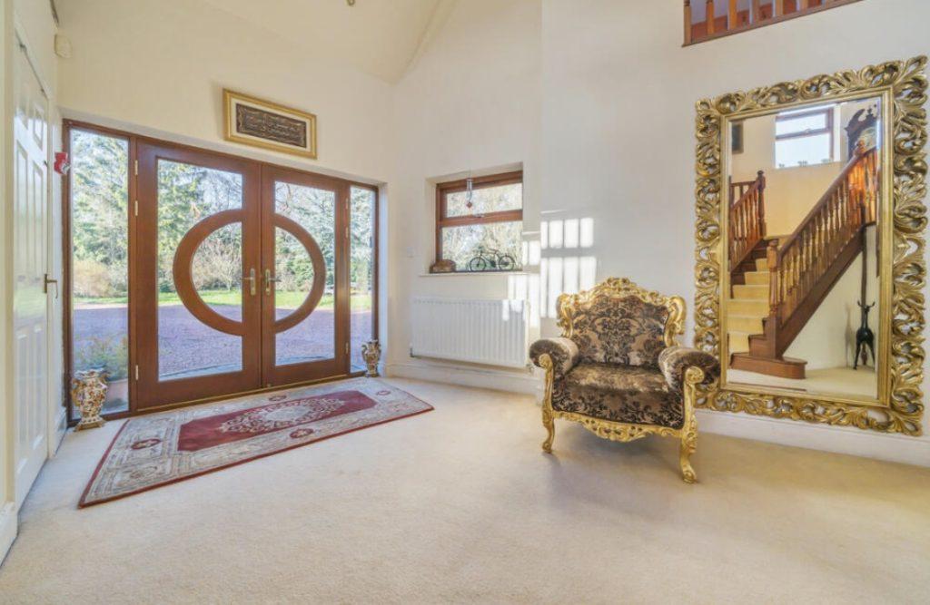Property for sale by Auction A spacious, sunlit foyer on Rowan Avenue with double wooden doors featuring glass inlays, a large ornate mirror reflecting a staircase, and an elaborate armchair. A red and beige rug lies in front of the door. A small window and a decorative frame adorn the white walls. Presented by Palace Auctions