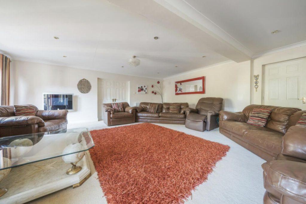 Property for sale by Auction A spacious living room on Rowan Avenue features multiple brown leather sofas and armchairs, a large rectangular red shaggy rug covering the light-colored floor, and a glass coffee table with a beige base. The walls are adorned with a large red-framed mirror and various wall decor. Presented by Palace Auctions