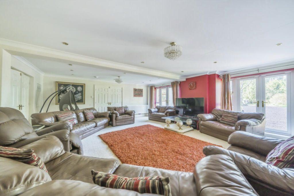 Property for sale by Auction Spacious living room on Rowan Avenue featuring multiple brown leather sofas arranged around a large orange rug. The room includes a flat-screen TV, large windows for natural light, and modern decor accents. A chandelier hangs from the ceiling, adding an elegant touch. Presented by Palace Auctions