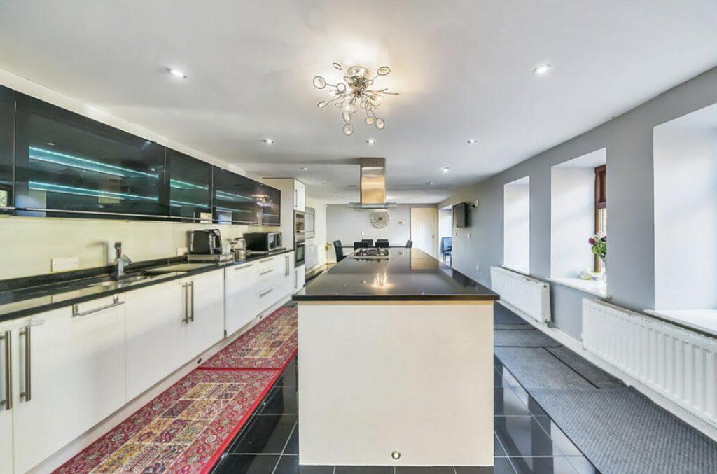 Property for sale by Auction A modern kitchen on Rowan Avenue features a long island counter with a sleek black countertop and white cabinets. Overhead lighting includes a unique chandelier, while stainless steel appliances line the left wall. Large windows and radiators adorn the right, with a colorful runner rug adding warmth. Presented by Palace Auctions