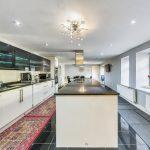 Property for sale by Auction A modern kitchen on Rowan Avenue features a long island counter with a sleek black countertop and white cabinets. Overhead lighting includes a unique chandelier, while stainless steel appliances line the left wall. Large windows and radiators adorn the right, with a colorful runner rug adding warmth. Presented by Palace Auctions