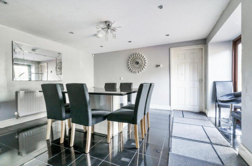 Property for sale by Auction A modern dining area on Rowan Avenue features a glass table with six upholstered chairs on a shiny black tiled floor. The light gray walls boast a sunburst wall decoration and a large mirror, while recessed lighting and a unique ceiling light fixture ensure the room is well-lit. Presented by Palace Auctions