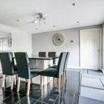 Property for sale by Auction A modern dining area on Rowan Avenue features a glass table with six upholstered chairs on a shiny black tiled floor. The light gray walls boast a sunburst wall decoration and a large mirror, while recessed lighting and a unique ceiling light fixture ensure the room is well-lit. Presented by Palace Auctions