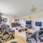 Property for sale by Auction A spacious living room on Rowan Avenue with patterned black and gold sofas, a large flat-screen TV, and a glass coffee table. The room features chandeliers, ornate curtains, and light-colored walls. A round mirror hangs on a wall near a large window, allowing natural light to enter. Presented by Palace Auctions