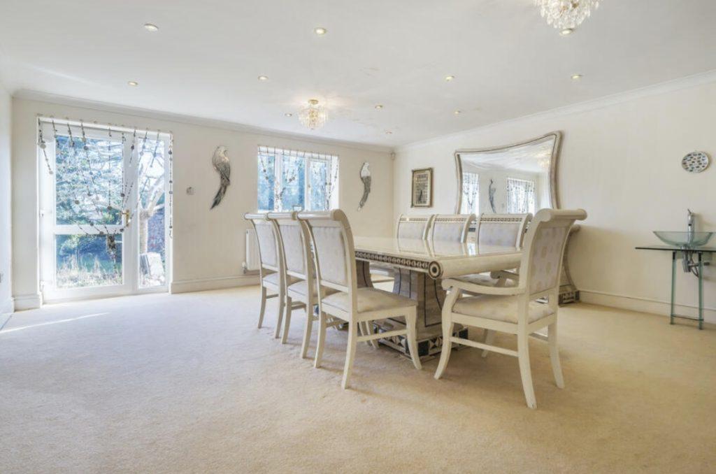 Property for sale by Auction A spacious dining room on Rowan Avenue boasts a long dining table surrounded by eight elegant chairs. The room features large windows with sheer curtains, a chandelier, wall decorations, a large mirror, and a wall clock. The carpeted floor adds warmth, while a minimalist console table sits in the corner. Presented by Palace Auctions