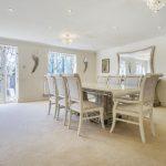 Property for sale by Auction A spacious dining room on Rowan Avenue boasts a long dining table surrounded by eight elegant chairs. The room features large windows with sheer curtains, a chandelier, wall decorations, a large mirror, and a wall clock. The carpeted floor adds warmth, while a minimalist console table sits in the corner. Presented by Palace Auctions