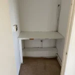 Property for sale by Auction A small, empty room with white walls and a tan carpet at 159 Collingwood Court, Washington. There is a built-in desk with peeling paint and visible wear attached to the back wall. A light switch is mounted on the left wall, and some exposed pipes run vertically along the back wall. Presented by Palace Auctions