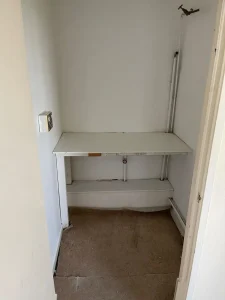 Property for sale by Auction A small, empty room with white walls and a tan carpet at 159 Collingwood Court, Washington. There is a built-in desk with peeling paint and visible wear attached to the back wall. A light switch is mounted on the left wall, and some exposed pipes run vertically along the back wall. Presented by Palace Auctions