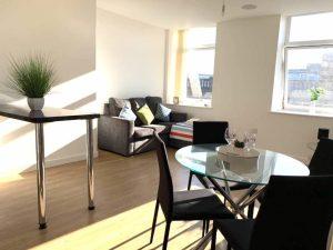 Property for sale by Auction A modern, brightly lit living area features a gray couch with colorful cushions, a round glass dining table with black chairs, and a tall table with a potted plant. Large windows allow natural light to flood the hardwood floor, creating a warm and inviting atmosphere. Sold by Palace Auctions. Presented by Palace Auctions