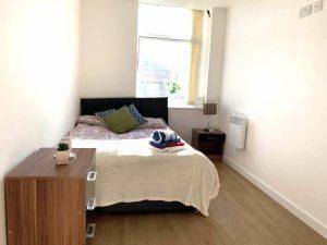 Property for sale by Auction A small, minimally furnished bedroom sold by Palace Auctions features a single bed against the far wall, covered in a white blanket with a folded blanket and pillow on top. A wooden bedside table with a plant is on the left, and a window with a partial view of the outdoors is in the background. Presented by Palace Auctions