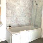 Property for sale by Auction A bathroom with a white bathtub featuring a glass shower screen and grey tiled walls. A white towel hangs over the side of the bathtub. There is a white pedestal sink with a small plant on the counter. Sold by Palace Auctions, a toilet is partially visible on the right. Presented by Palace Auctions