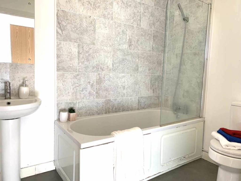 Property for sale by Auction A bathroom with a white bathtub featuring a glass shower screen and grey tiled walls. A white towel hangs over the side of the bathtub. There is a white pedestal sink with a small plant on the counter. Sold by Palace Auctions, a toilet is partially visible on the right. Presented by Palace Auctions