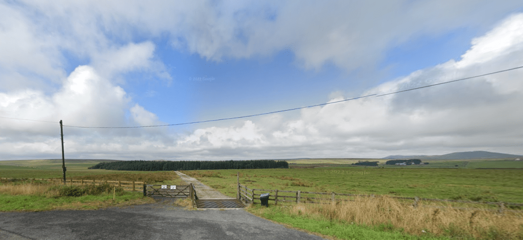 Property for sale by Auction A rural landscape with a narrow asphalt road leading through fields of green grass towards a dense forest. In the foreground, a cattle grid and a wired fence are visible. With farmland buildings seen in the distance, it's easy to imagine the land at Bolventor sold amidst such scenic beauty. Presented by Palace Auctions