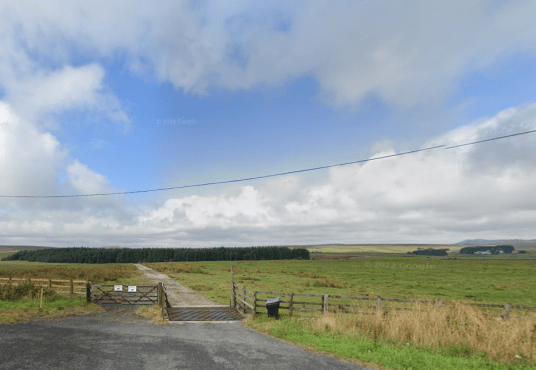 Property for sale by Auction A rural landscape with a narrow asphalt road leading through fields of green grass towards a dense forest. In the foreground, a cattle grid and a wired fence are visible. With farmland buildings seen in the distance, it's easy to imagine the land at Bolventor sold amidst such scenic beauty. Presented by Palace Auctions