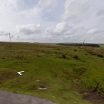 Property for sale by Auction A vast, open grassy landscape under a cloudy sky, with patches of green field stretching into the distance. A dirt road runs along the bottom edge, and a wire fence with wooden posts cuts across the scene. Recently sold land at Bolventor lies just beyond the directional arrow visible on the ground. Presented by Palace Auctions