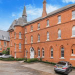 Property for sale by Auction A red-brick building with gothic-style windows and a tall spire stands under a clear blue sky. Several cars are parked in front on a paved driveway. Another similar building, partly visible on the left side and surrounded by trees and bushes, was previously marketed by Palace Auctions. Presented by Palace Auctions