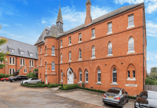 Property for sale by Auction A red-brick building with gothic-style windows and a tall spire stands under a clear blue sky. Several cars are parked in front on a paved driveway. Another similar building, partly visible on the left side and surrounded by trees and bushes, was previously marketed by Palace Auctions. Presented by Palace Auctions